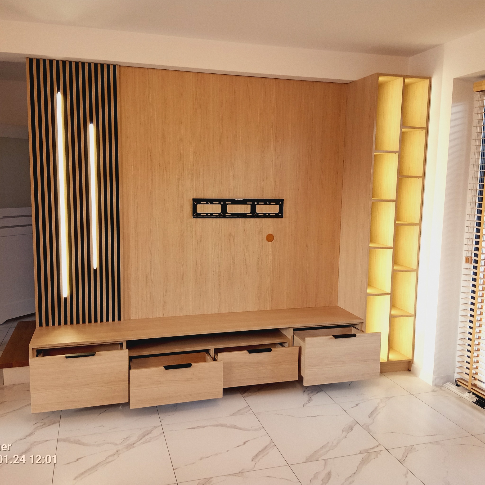 Drewniana zabudowa ściany z szafkami i półkami, podświetlona listwami LED. Na ścianie uchwyt na telewizor. Otwarta szuflada. Marmurowe płytki podłogowe.