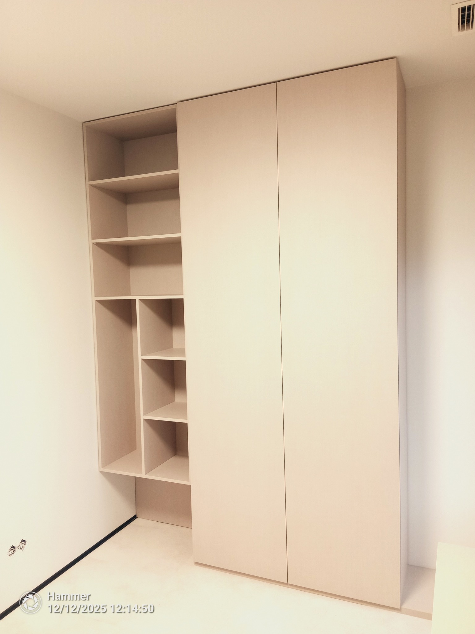 Jasna, minimalistyczna szafa wnękowa z otwartymi półkami po lewej stronie i zamkniętymi drzwiami po prawej, w nowym wnętrzu. Widoczne gniazdka elektryczne.