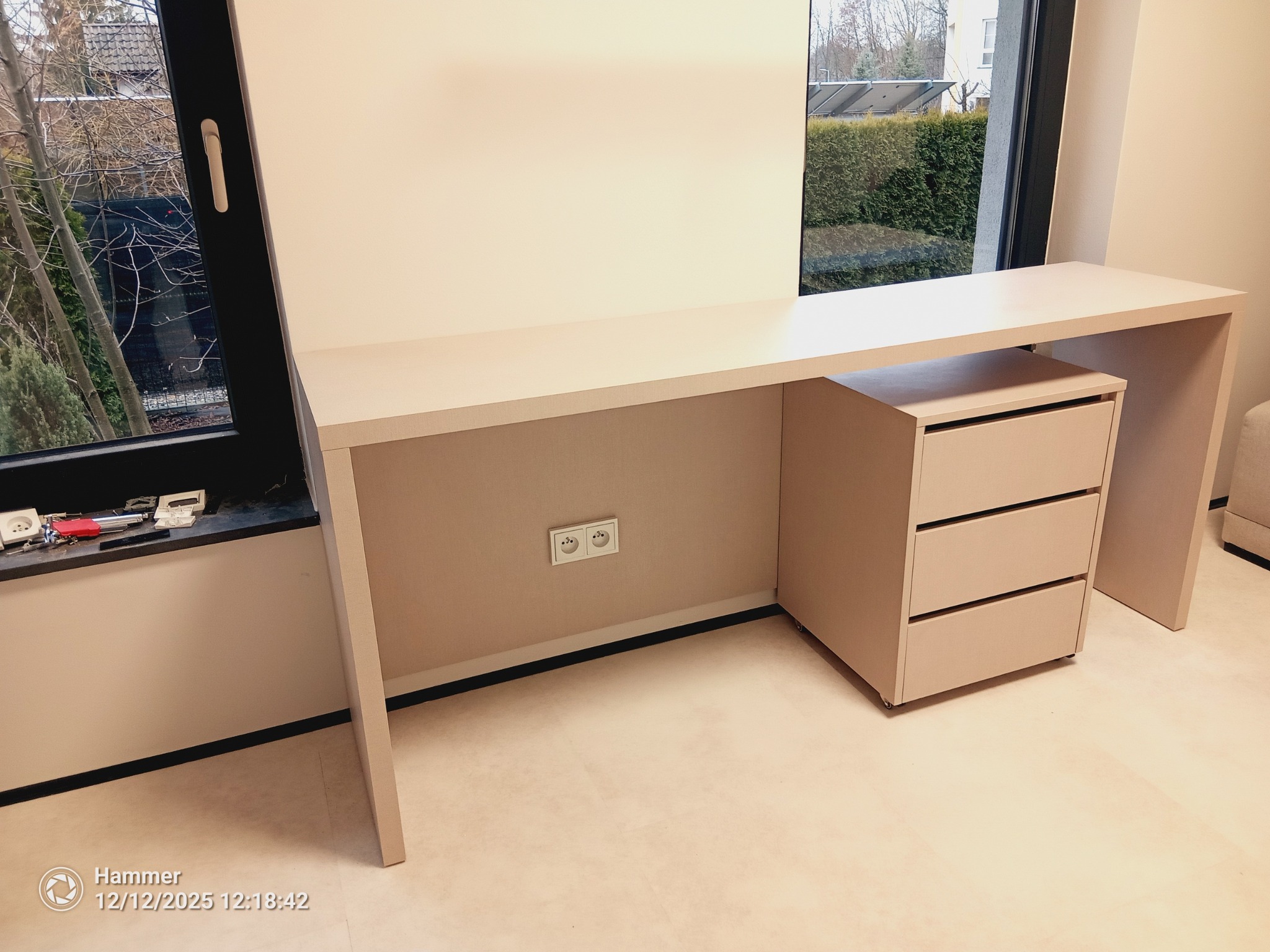 Beżowe biurko na wymiar z trzema szufladami, ustawione przy oknie. Na parapecie widoczne narzędzia. Minimalistyczny design, jasne wnętrze.