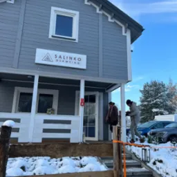 Szary, drewniany dom z białymi detalami i szyldem 'Salinko Glamping' w zimowej scenerii, z widocznymi schodami wejściowymi w trakcie budowy i dwoma osobami w pobliżu wejścia.