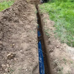 Długi, wąski wykop w ziemi z niebieską folią wewnątrz, z mini koparką w tle, obok przyczepa kempingowa.