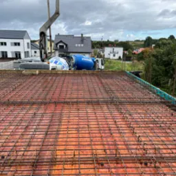 Zbrojenie stropu przygotowane do zalewania betonem, widoczna pompa do betonu i betoniarka w tle, zabudowa jednorodzinna w oddali, pochmurne niebo.