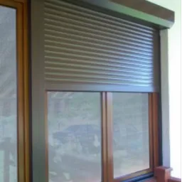 Częściowo zasunięta brązowa osłona okienna zewnętrzna, widoczna z zewnątrz budynku, z okiennicami siatkowymi w tle.