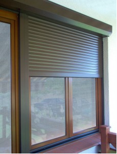 Częściowo zasunięta brązowa osłona okienna zewnętrzna, widoczna z zewnątrz budynku, z okiennicami siatkowymi w tle.