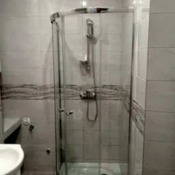 Szklana kabina prysznicowa narożna z chromowanymi detalami, umieszczona w łazience wyłożonej jasnymi płytkami z dekoracyjnym pasem, widoczna umywalka z szafką