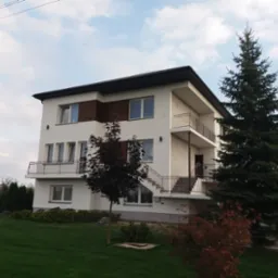 Trzykondygnacyjny dom z białymi ścianami, brązowymi akcentami i balkonami, otoczony zielonym trawnikiem i drzewami iglastymi oraz liściastymi.