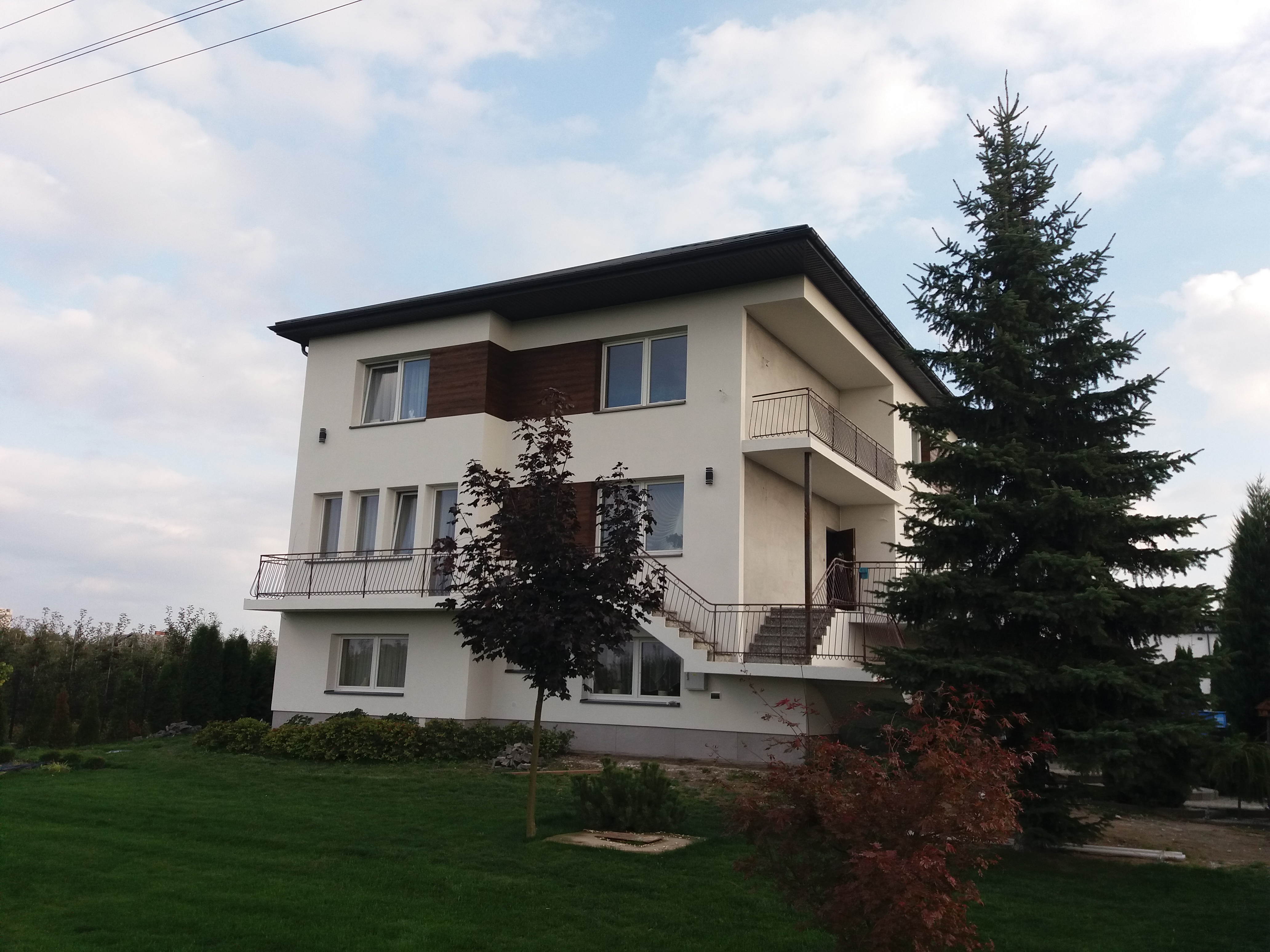 Trzykondygnacyjny dom z białymi ścianami, brązowymi akcentami i balkonami, otoczony zielonym trawnikiem i drzewami iglastymi oraz liściastymi.