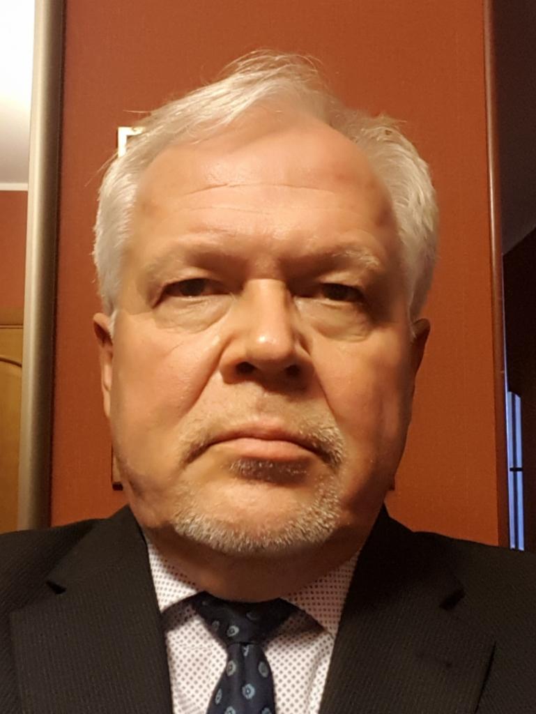 Portret mężczyzny w garniturze i krawacie z niebieskimi wzorami, na tle pomarańczowej ściany.