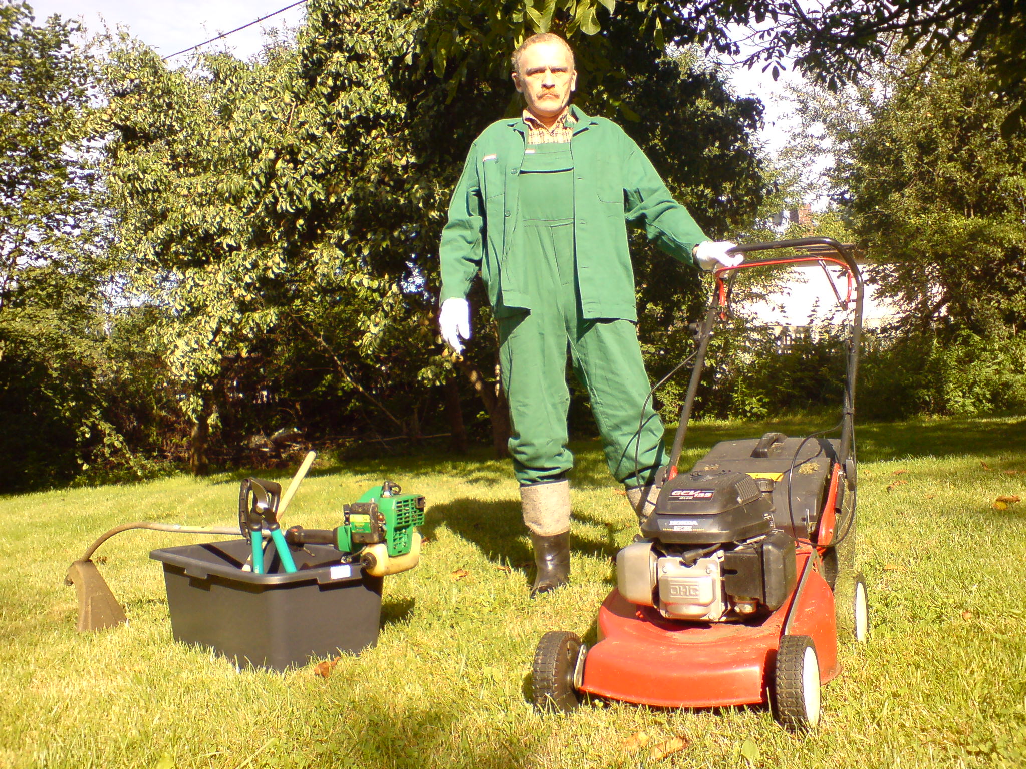 Mężczyzna w zielonym kombinezonie i białych rękawicach stoi na trawniku obok czerwonej kosiarki, pojemnika z narzędziami ogrodniczymi i podkaszarki.
