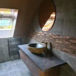 Nowoczesna łazienka z kamienną umywalką, baterią w kolorze złota i owalnym lustrem. Na ścianie dekoracyjna mozaika. Widoczne okno dachowe.