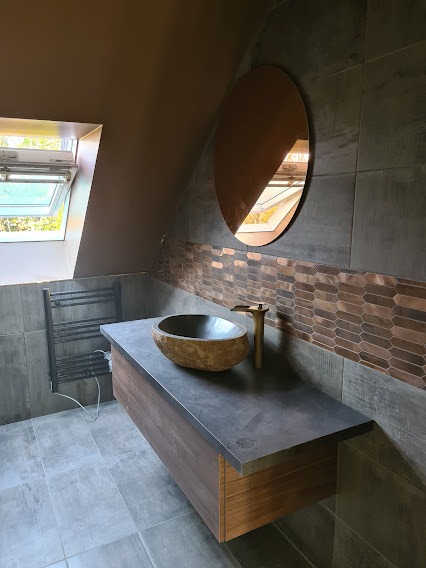 Nowoczesna łazienka z kamienną umywalką, baterią w kolorze złota i owalnym lustrem. Na ścianie dekoracyjna mozaika. Widoczne okno dachowe.