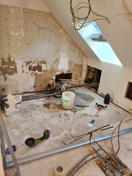 Adaptacja poddasza: ściana z ubytkami tynku, widoczne instalacje, narzędzia i materiały budowlane. Okno dachowe w skośnym suficie. Prace w toku.