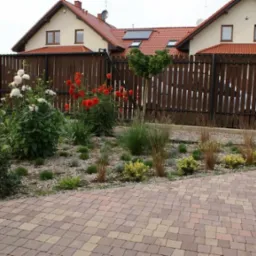 Nowoczesny ogr&oacute;d przydomowy z rabatami żwirowymi, ozdobnymi trawami, kwitnącymi daliami i wybrukowanym podjazdem na tle drewnianego ogrodzenia.