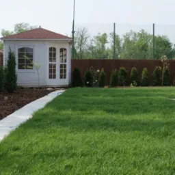 Świeżo założony trawnik z rolki, widoczny biały domek ogrodowy z czerwonym dachem, obrzeże z jasnych płyt betonowych i nasadzenia z tuj.