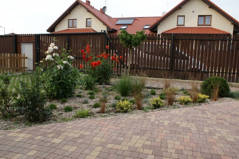 Nowoczesny ogród przydomowy z rabatami żwirowymi, ozdobnymi trawami, kwitnącymi daliami i wybrukowanym podjazdem na tle drewnianego ogrodzenia.