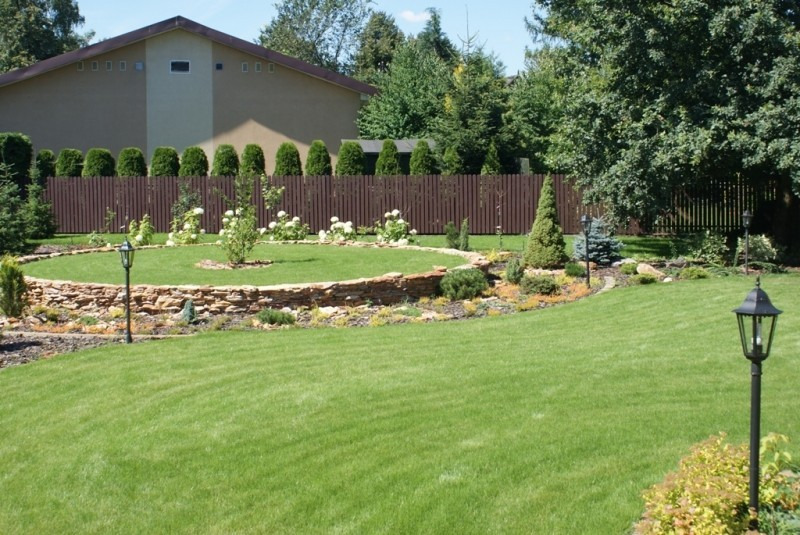 Utrzymany w idealnym stanie trawnik z równo skoszoną trawą, otoczony niskim murkiem z kamienia, z widocznymi lampami ogrodowymi i dekoracyjnymi krzewami, na tle brązowego ogrodzenia i domu.