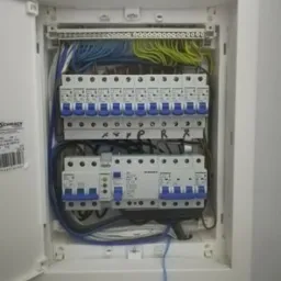 Otwarta skrzynka elektryczna z bezpiecznikami, widoczne okablowanie w kolorach niebieskim, zielonym i białym, oraz oznaczenia na listwach zaciskowych.