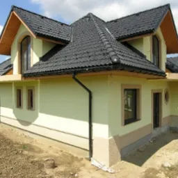 Dwukondygnacyjny dom z czarną dachówką, jasną elewacją i dekoracyjnymi elementami z piaskowca, widoczny z boku na działce z przygotowanym terenem.