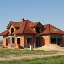 Surowy stan domu z czerwonej cegły, z dachem z czerwonej dachówki, widoczny stos piasku przed budynkiem i niezabudowane otwory okienne, w tle zieleń i błękitne niebo.