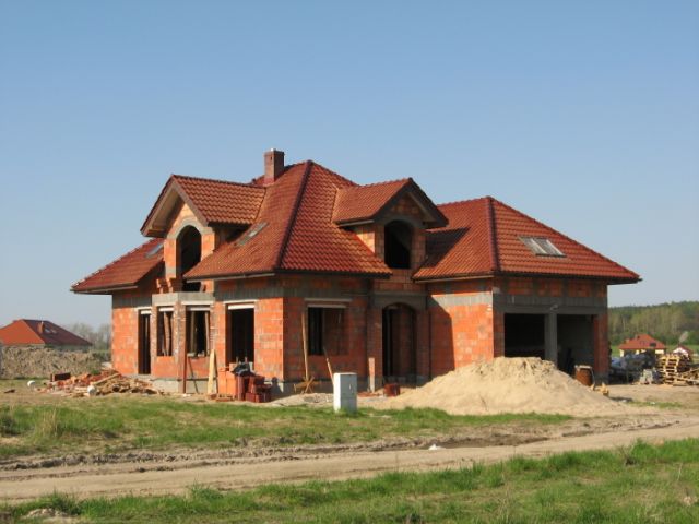 Surowy stan domu z czerwonej cegły, z dachem z czerwonej dachówki, widoczny stos piasku przed budynkiem i niezabudowane otwory okienne, w tle zieleń i błękitne niebo.