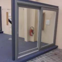 Szare aluminiowe okno przesuwne z widocznym wnętrzem pomieszczenia, w którym stoi gaśnica, na podłodze wzór szachownicy.