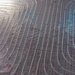 Ułożone, białe rury ogrzewania podłogowego na folii z czerwoną siatką. Widoczne czarne uchwyty mocujące rury do podłoża. Instalacja przed wylaniem posadzki.