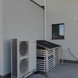 Zewnętrzna jednostka pompy ciepła Panasonic Aquarea obok drewnianej osłony z czarnym dachem, przy białej ścianie domu z dwoma oknami i czarną rynną.