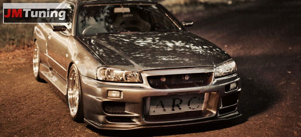 Szary samochód sportowy Nissan Skyline R34 z widocznym logo JMTuning w lewym górnym rogu, obniżonym zawieszeniem i zmodyfikowanym przednim zderzakiem z napisem ARC.