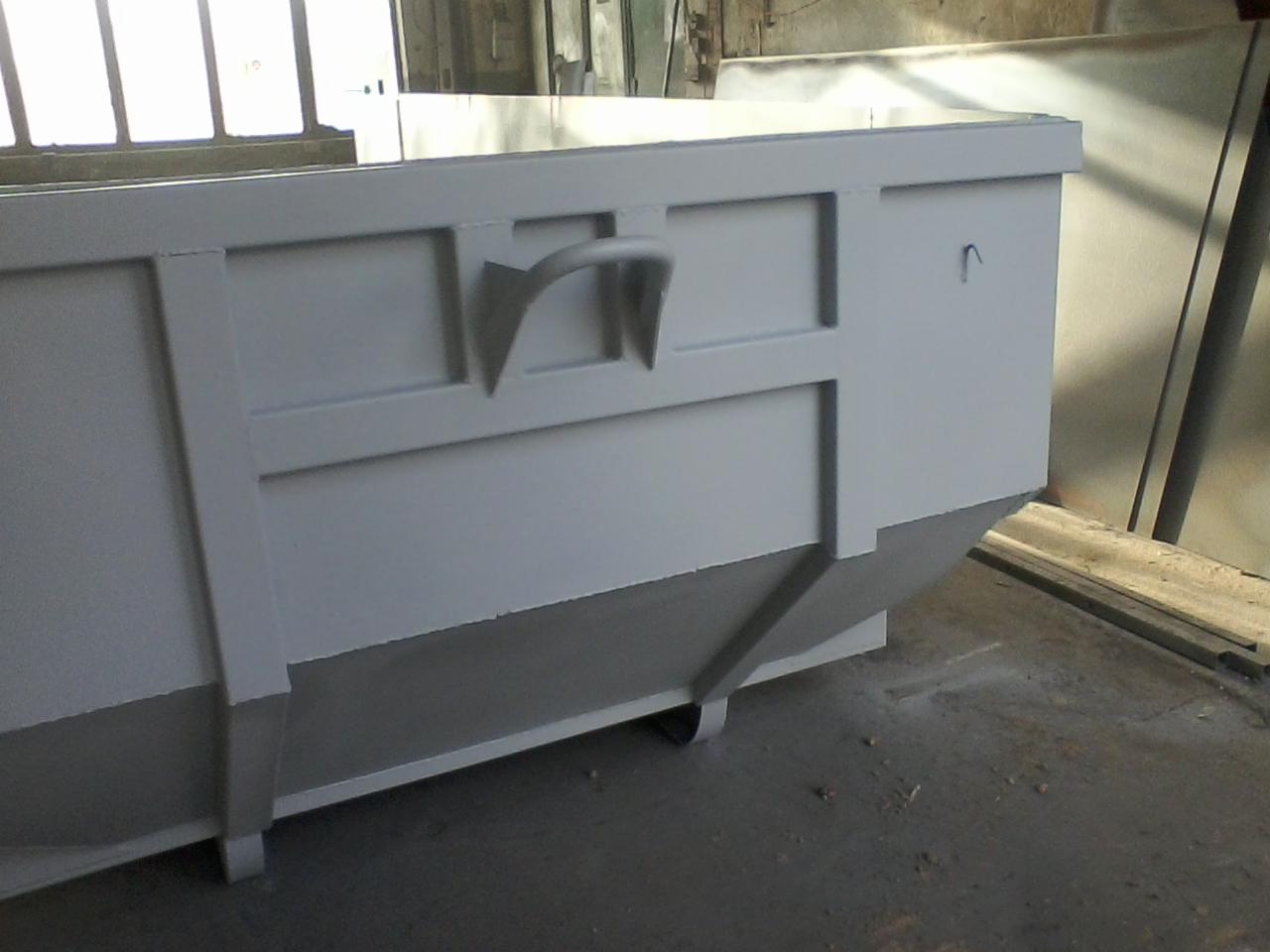 Metalowy kontener pomalowany na szaro, z widocznymi uchwytami i elementami konstrukcyjnymi, ustawiony na betonowej posadzce w hali.