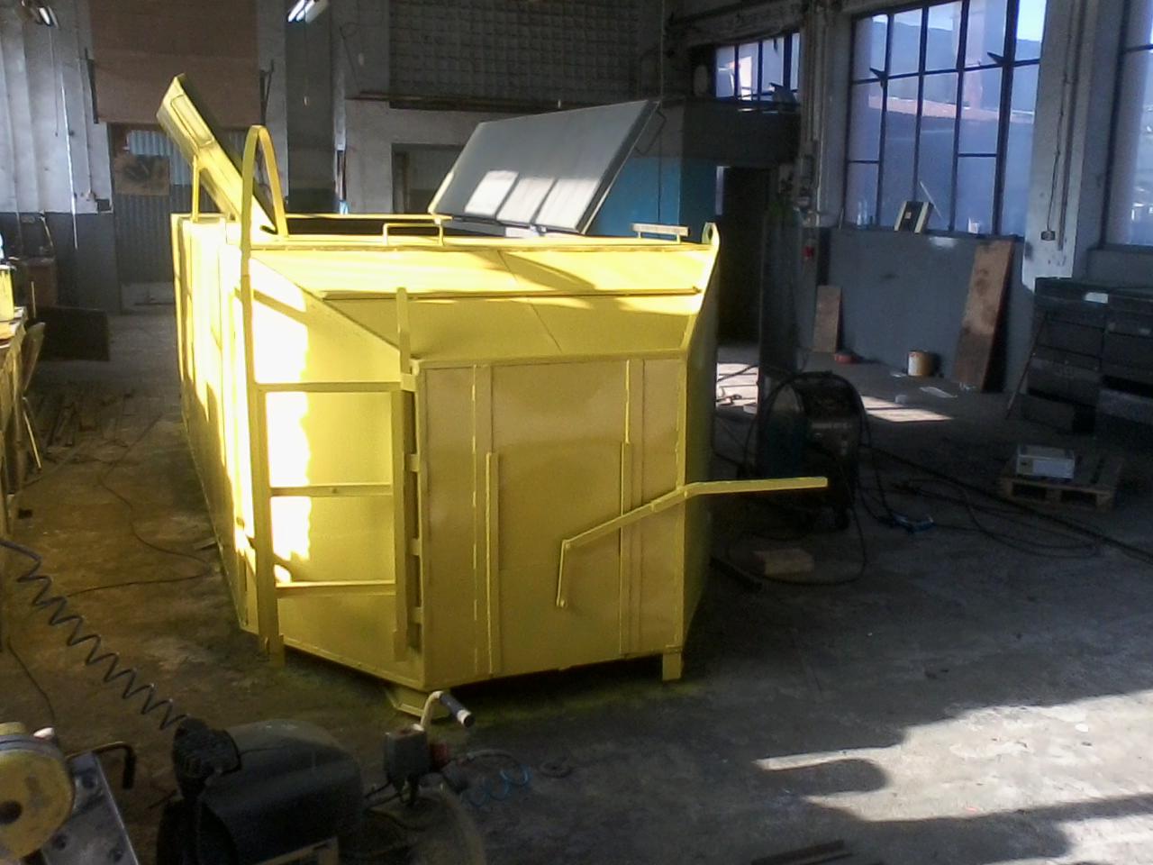 Żółta, metalowa konstrukcja przypominająca kontener z otwartą klapą, widoczna w hali produkcyjnej z betonową podłogą i dużymi oknami w tle.