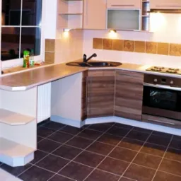 Kąt ujęcia na nowoczesną kuchnię z zabudową w odcieniach beżu i brązu, z czarnym zlewem i płytą grzewczą, podświetlona punktowymi lampami, z widocznym oknem i elementami dekoracyjnymi na ścianie.