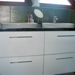 Biała szafka łazienkowa pod umywalki nablatowe z czarnym blatem, srebrne uchwyty, tło stanowi błyszcząca mozaika w odcieniach szarości.