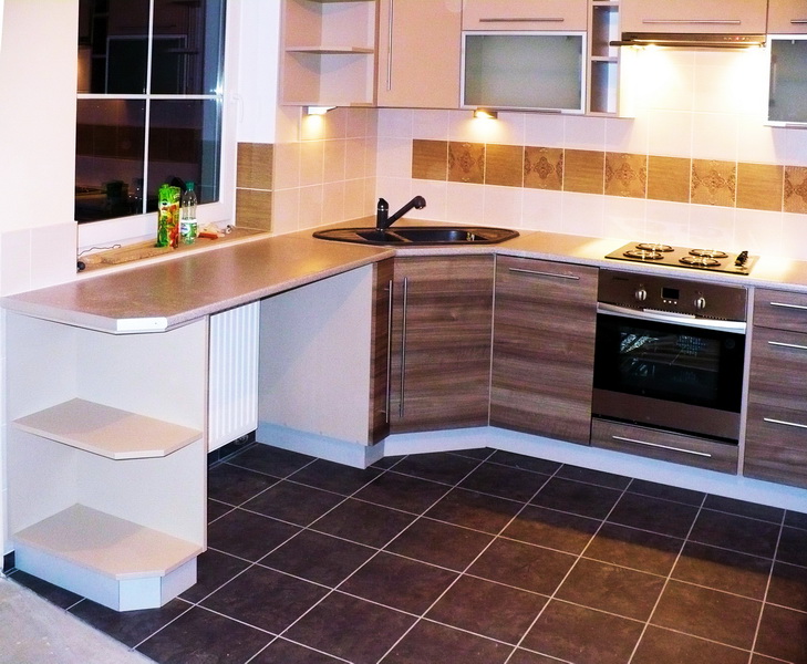 Kąt ujęcia na nowoczesną kuchnię z zabudową w odcieniach beżu i brązu, z czarnym zlewem i płytą grzewczą, podświetlona punktowymi lampami, z widocznym oknem i elementami dekoracyjnymi na ścianie.