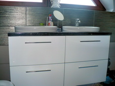 Biała szafka łazienkowa pod umywalki nablatowe z czarnym blatem, srebrne uchwyty, tło stanowi błyszcząca mozaika w odcieniach szarości.