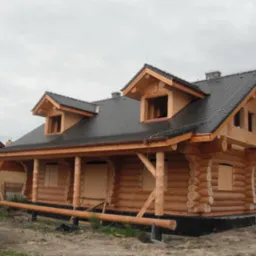 Surowy drewniany dom z bali, z czarnym dachem z dachówki bitumicznej, na etapie budowy, z deskami zakrywającymi otwory okienne, widok z zewnątrz w pochmurny dzień.