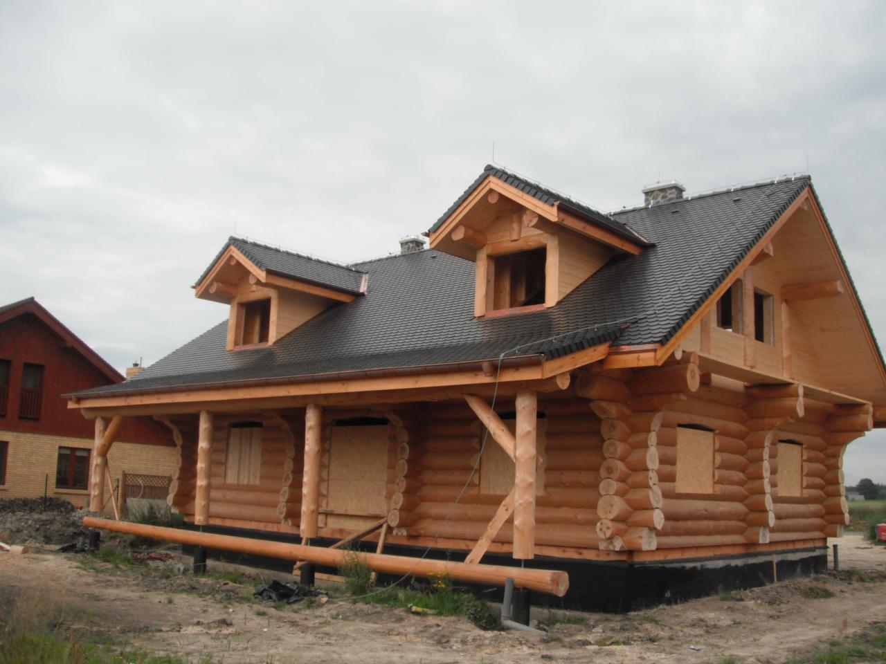 Surowy drewniany dom z bali, z czarnym dachem z dachówki bitumicznej, na etapie budowy, z deskami zakrywającymi otwory okienne, widok z zewnątrz w pochmurny dzień.
