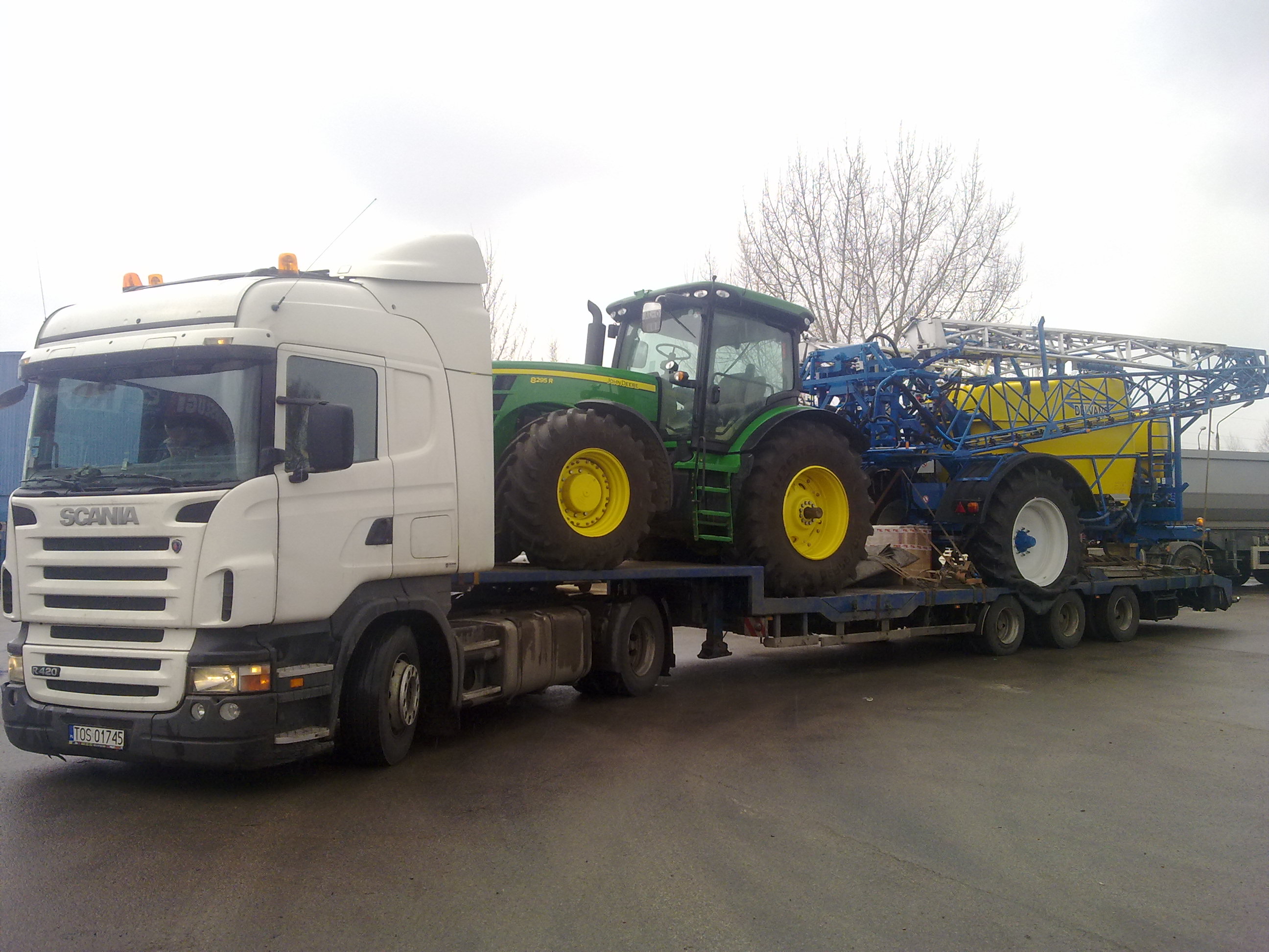 Ciężarówka Scania R420 z naczepą niskopodwoziową przewożąca zielony traktor John Deere 8295R i opryskiwacz polowy z żółtym zbiornikiem na tle szarego nieba.