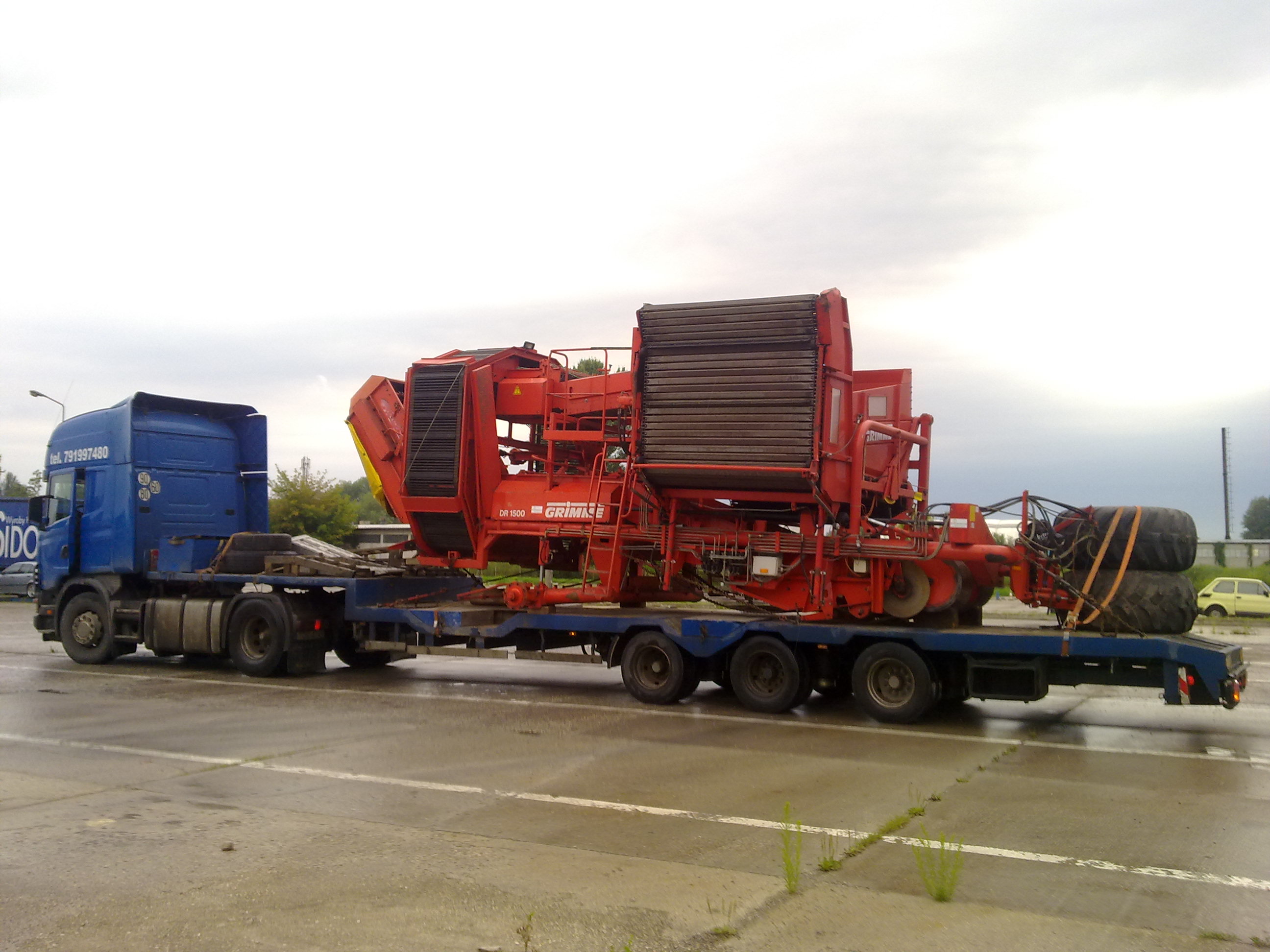 Niebieska ciężarówka z naczepą przewozi czerwoną maszynę rolniczą Grimme DR 1500, z dodatkowymi oponami zabezpieczonymi pasami na naczepie, na mokrym placu.