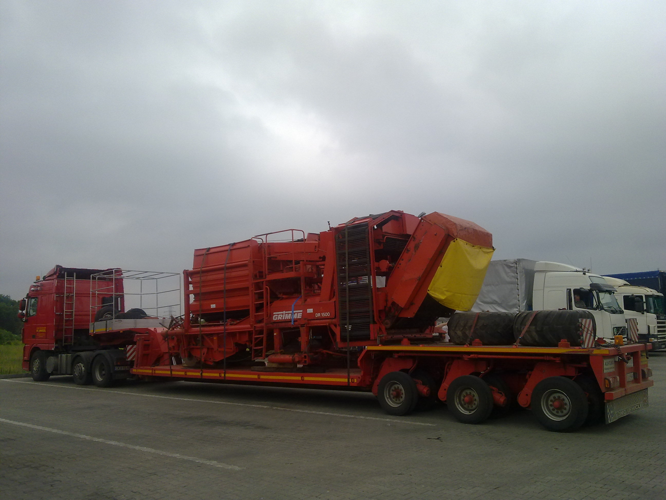 Czerwona ciężarówka niskopodwoziowa przewozi dużą, czerwoną maszynę rolniczą Grimme DR 1500 z żółtą plandeką zabezpieczającą, na parkingu w pochmurny dzień. W tle zaparkowane inne ciężarówki.