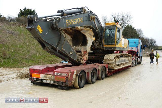 Żółto-szara koparka marki Ennstone przewożona na czerwonej niskopodwoziowej platformie transportowej, stojącej na błotnistym terenie; w tle widoczni dwaj mężczyźni i częściowo błękitna ciężarówka.