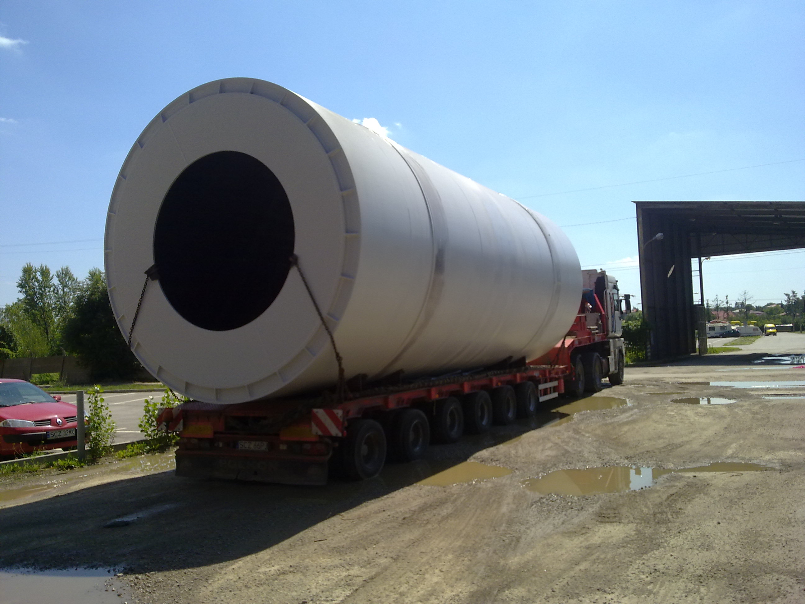 Nietypowy ładunek: Duży, biały, cylindryczny element z otworem w środku, zabezpieczony łańcuchami, przewożony na niskopodwoziowej naczepie ciężarowej na tle pochmurnego nieba i kałuż na placu.