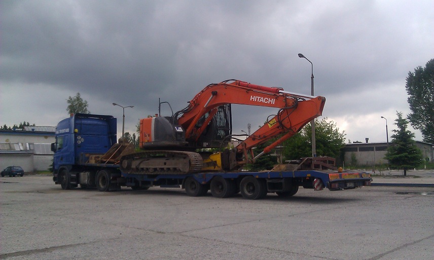 Niebieska ciężarówka z pomarańczową koparką Hitachi na niskopodwoziowej platformie, transportowana na tle pochmurnego nieba.