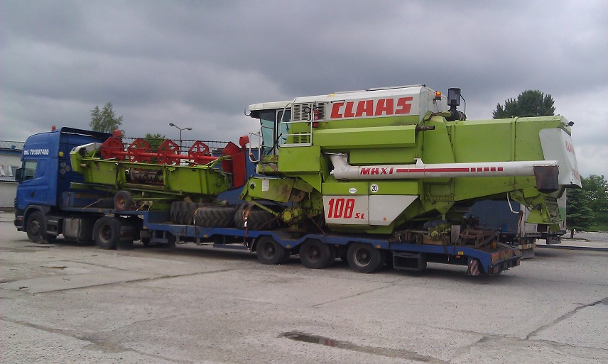 Niebieska ciężarówka z naczepą przewozi kombajn rolniczy Claas Maxi 108 SL na betonowym placu w pochmurny dzień.