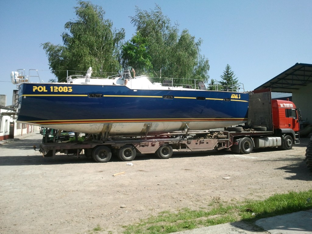 Transport jachtu ADAL 3 na niskopodwoziowej platformie, widoczny numer rejestracyjny POL 12083, czerwona kabina ciężarówki, drzewa w tle.