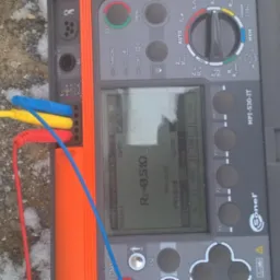 Miernik Sonel MPI-530-IT z podłączonymi przewodami (czerwony, ż&oacute;łty, niebieski) wyświetlający wynik pomiaru rezystancji uziemienia na ekranie LCD, leżący na ziemi z grudkami śniegu.