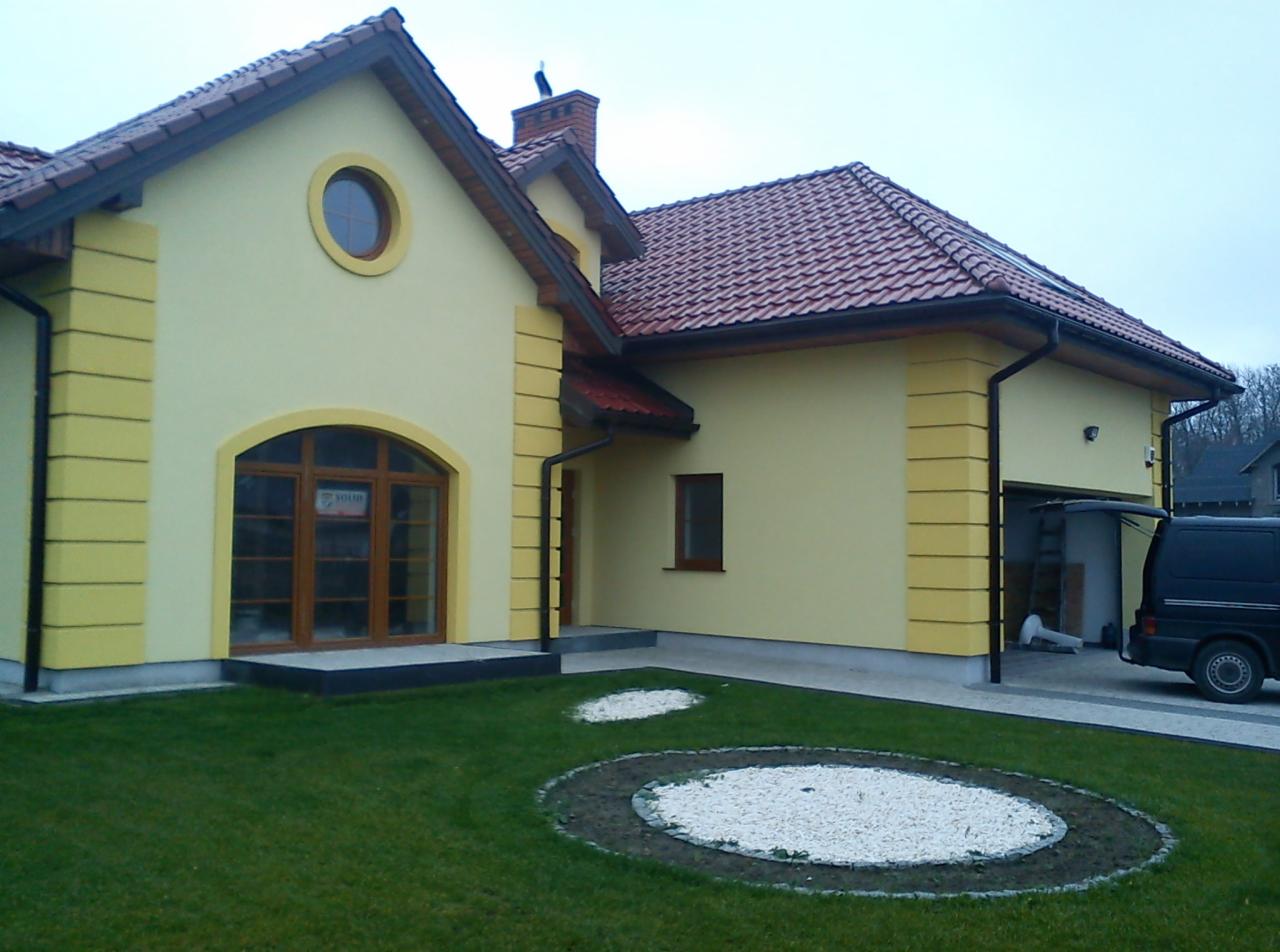 Żółta elewacja domu jednorodzinnego z dekoracyjnymi detalami, widok z ogrodu, boczny. Czerwona dachówka, brązowe okna. Na dachu siedzi ptak.