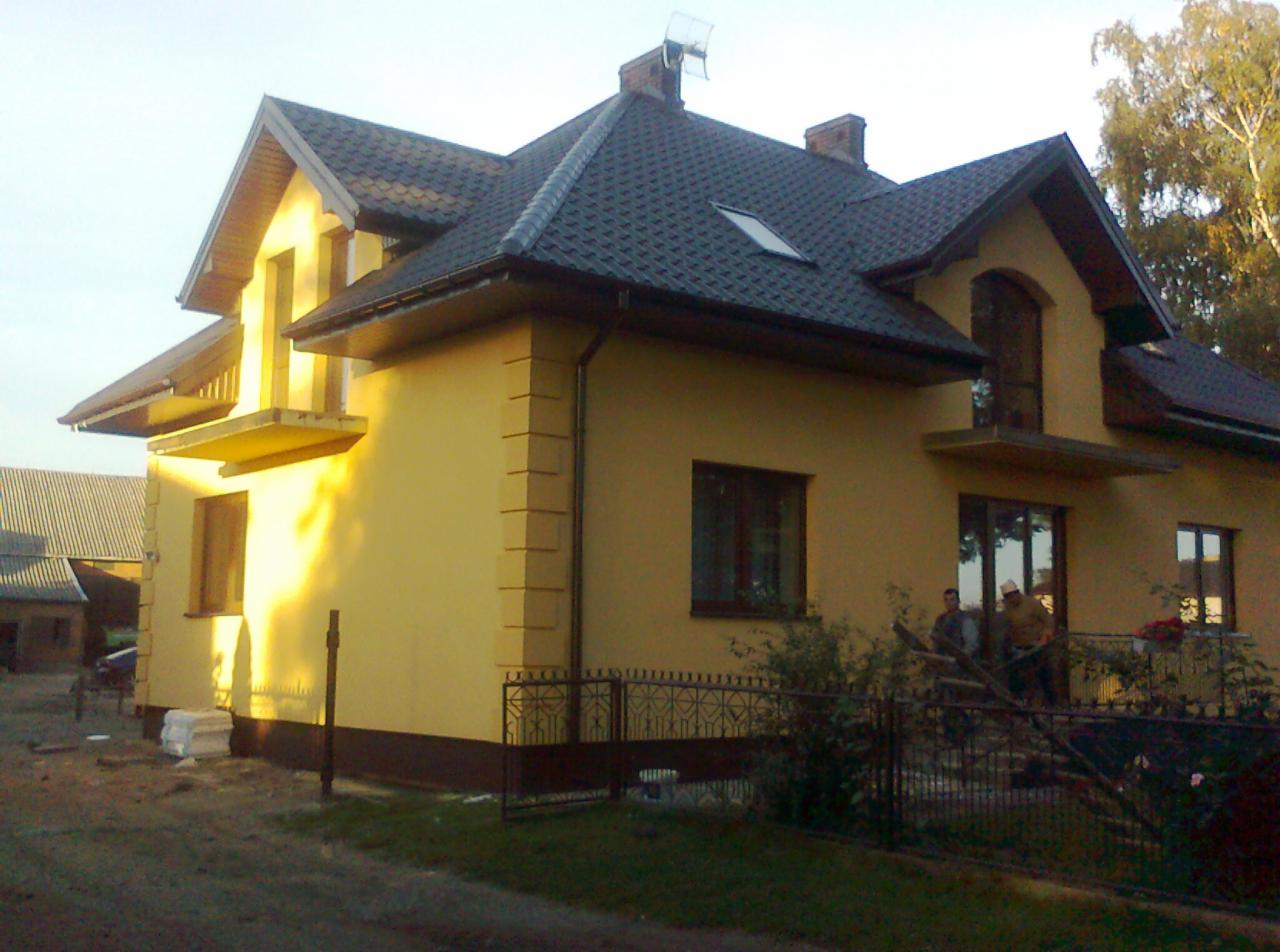 Żółty dom jednorodzinny z ciemną dachówką, widok z boku, z elementami dekoracyjnymi na elewacji i anteną na dachu. Częściowo ogrodzony, z widoczną zielenią.