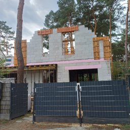 USŁUGI REMONTOWO BUDOWLANE - Budowa domu z betonu komórkowego w lesie, widoczne drewniane szalunki okien i różowa izolacja. W tle wysokie drzewa i fragment ogrodzenia.