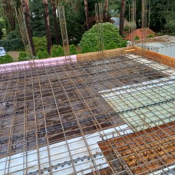 USŁUGI REMONTOWO BUDOWLANE - Zbrojenie stropu z widocznymi prętami, siatką i elementami izolacji termicznej na tle drzew. Widok z góry ukazuje etap budowy domu.