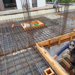 USŁUGI REMONTOWO BUDOWLANE - Zbrojenie fundamentów z drewnianym szalunkiem, widoczne pręty zbrojeniowe i pracownik przy szalunku. W tle budynek z oknami i osoba siedząca na murku.